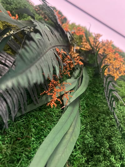 This is a close-up, high-angle shot of a decorative green moss wall with artificial tropical plants. The moss forms a dense, textured carpet covering the surface. Large, dark green palm-like leaves with distinct veins are prominent in the foreground, partially obscuring smaller, delicate orange flowers that are scattered across the moss. In the background, more of the orange flowers are visible, interspersed with green foliage, all mounted on a wall. The lighting suggests an indoor environment, possibly a commercial space or home decor installation, and there is no indication of any outdoor elements or specific location within the city of Минзаг, Russia. The scene is static, with no people or activity depicted.