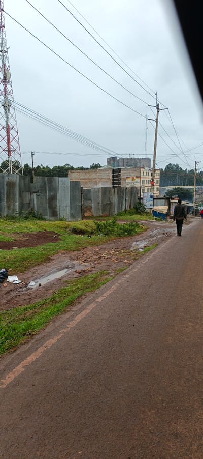 A view along a wet, paved road with grassy, muddy verges and trash. A corrugated metal fence obscures a building under construction. Power lines stretch overhead. A tall red and white communication tower stands in the distance, and a person walks down the road, away from the viewer. The sky is overcast. Another building can be seen in the distance, it has several floors and advertisements.