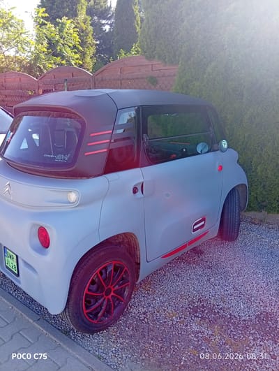 A light gray, two-door Citroën Ami electric car is parked on a gravel surface next to a paved walkway. The car has a black roof with red accents on the rear quarter panel and a red stripe along the lower body. The wheels have black rims with red accents. A license plate, "DWR JL28", is visible on the rear of the car. The car is situated outdoors, with a wooden fence and lush green trees in the background. The image is watermarked with "POCO C75" in the bottom left corner and a date and time stamp, "08.06.2026 08:31", in the bottom right corner. The scene appears to be located in Kiełczów, Poland, based on the license plate format and the overall setting.