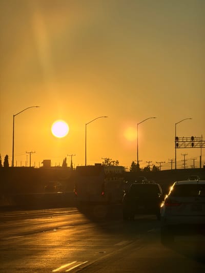 The image shows a sunset over a highway in Burbank, California, United States of America. The sun is low in the sky, casting a warm glow over the scene. Cars are visible on the highway, driving in both directions. Streetlights are also present, illuminating the road. In the background, there are buildings and trees. The overall impression is one of a peaceful evening commute.