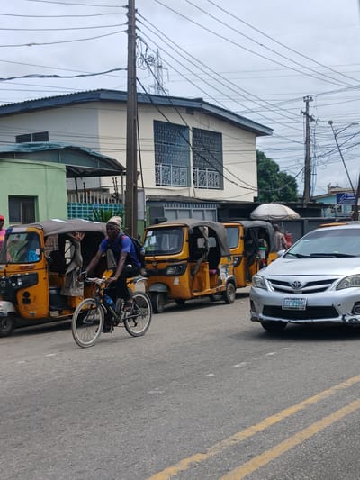 In Animashawun, Nigeria, a street scene unfolds under an overcast sky. A man wearing a blue t-shirt and a patterned head covering rides a bicycle on a paved road. He is positioned in front of two yellow auto-rickshaws, one of which has its door open. To the right of the auto-rickshaws, a silver sedan is parked on the road. Behind the vehicles, a light-colored building with blue-trimmed windows stands, and beyond that, more buildings and utility poles with numerous wires are visible. The road is marked with yellow lines.