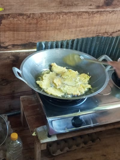 An overhead shot captures a close-up of battered food frying in a large, shallow metal wok on a gas stove. The wok is filled with bubbling oil, and several pieces of golden-brown batter-coated food are submerged. A metal spatula is used to turn the food. The wok rests on a two-burner gas stove with a silver-colored base and black knobs. The stove is situated on a wooden platform. Behind the stove, a wall constructed of horizontal wooden planks is visible. The background also includes a section of corrugated metal. In the lower left corner, part of a clear plastic bottle with a yellow cap is visible, along with a metal sieve and a piece of foam. In the lower right corner, an egg carton is visible. The scene suggests a domestic cooking environment in Turumame, Indonesia.
