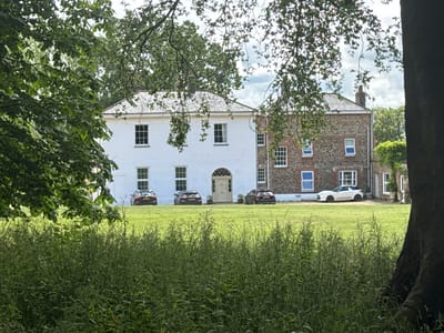 The image depicts a detached residential property in North Norfolk, United Kingdom. The house features a white facade on the left, with a section of stone and brick construction on the right. Several cars are parked on the manicured lawn in front of the building. The foreground is dominated by tall green grass and foliage, with a large tree trunk on the far right obscuring part of the scene. Overhead, branches and leaves from a deciduous tree frame the top of the image. The sky is partly cloudy. No people are visible.