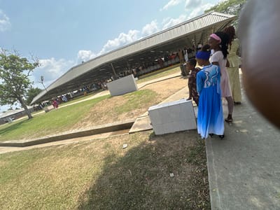 This image captures an outdoor scene in Ozuaha, Nigeria, under a partly cloudy blue sky. In the foreground, a dry grassy area transitions to a concrete drainage channel running horizontally, followed by a paved concrete edge or walkway along the right side of the frame.
Four individuals, primarily seen from behind, stand on this concrete edge, facing towards the left midground. The closest individual on the right wears a long, flowing blue and white gradient dress with a wave-like pattern. To their immediate left, a shorter individual wears a brown patterned shirt and a blue head covering. Adjacent to the individual in the blue dress, a young female is visible, wearing a light pink top, white leggings, and a pink headband, with dark hair tied back. Further to the right, partially obscured by an out-of-focus element, another young female wears a light yellow/cream traditional-style long dress with textured fabric. A rectangular object, tiled in white, rests on the ground next to the paved edge near these individuals.
In the midground, a long, open-sided covered structure with a sloped, light-colored metal roof and visible support beams spans across the scene. Underneath this shelter, a large congregation of people is visible, seated in rows of chairs and standing, indicating a public gathering or event. A street light pole stands on the grassy area to the left of the covered structure. The ground underfoot is a mix of dry grass and bare earth. Trees are present in the far left background.