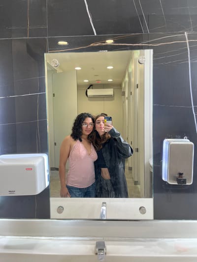 This image captures two young women taking a mirror selfie in a modern public restroom, likely located in San Buenaventura, Mexico.

The setting features stylish dark walls, possibly made of marble or granite, adorned with distinct white and light brown veining. A large, rectangular mirror dominates the center, reflecting the subjects and the restroom's interior. Mounted above the mirror is a metallic soap dispenser with a black top. Below the mirror, a white hand dryer with the brand "KAYSER" and a red logo is visible. The reflection in the mirror reveals a light-colored ceiling with several recessed circular lights and a white rectangular air unit on the far wall.

The two women are the primary subjects. The woman closer to the mirror's surface (on the left in the reflection) has curly dark hair, wears glasses, a light pink lace-trimmed tank top, and blue jeans. She displays a soft smile. The second woman, holding a smartphone with a light-colored case to capture the selfie, has long brown hair and is dressed in a black leather-like jacket over a dark top. She is making a playful "duck face" or pout for the camera. Both appear to be in good spirits.

The activity is a casual mirror selfie, suggesting a moment of friendship or fun. The lighting is artificial, consistent with an indoor setting. The image is rotated 90 degrees counter-clockwise.