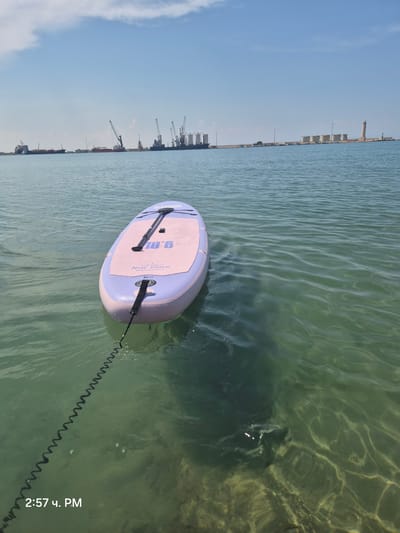 The image depicts a stand-up paddleboard floating on clear, turquoise water, tethered by a coiled leash. The paddleboard is light purple and pink, with "10'6" and "New Dawn" visible on its surface. In the background, a hazy industrial port with cranes and silos stretches along the horizon under a bright blue sky with scattered clouds. The scene is likely located near the port of Barletta, Italy, given the industrial waterfront and lighthouse visible in the distance. The water is calm with gentle ripples, and sunlight reflects off its surface, illuminating the sandy bottom below. There are no people visible in the image.