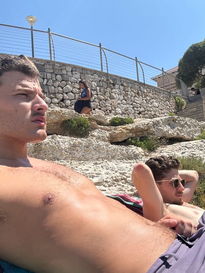 The image captures an outdoor scene on a clear, sunny day in a rocky coastal area of Nardò, Italy. In the foreground, an adult male lies shirtless on light-colored, uneven rock formations, with his head turned to the right and eyes partially closed from sunlight. His upper torso is prominent. To his right, another adult male reclines on a patterned towel, wearing sunglasses and with his hands behind his head. Both appear to be sunbathing.

In the midground, a stone retaining wall extends horizontally, topped by a metal railing with multiple curved supports and a single lamp post on the left. Below this wall, a female subject in a blue top and dark shorts is observed standing or crouching on the rocks, looking downward at an object held in her hands. To the far right of the scene, a series of stone steps ascends, with a partially visible male subject seated on them. Sparse green vegetation grows among the rocks. A building with a visible roofline is situated beyond the stone wall and steps on the right. The sky is uniformly blue and cloudless.