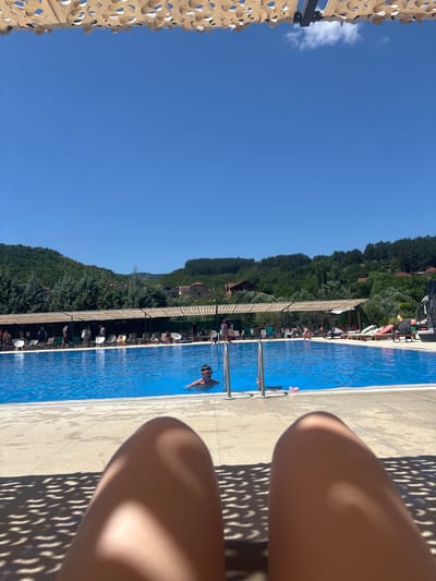 The image depicts an outdoor swimming pool on a sunny day. In the foreground, partially visible from a first-person perspective, are the legs of a person resting, with shadows from a patterned canopy above them. The pool itself is a large, bright blue expanse, with several people scattered around its edges and in the water.
A young boy wearing goggles is swimming in the pool, with his head and shoulders visible. Other individuals are seen sitting on lounge chairs or standing by the pool, some beneath shaded awnings. Beyond the pool area, the landscape is characterized by lush green hills and trees, with scattered houses visible on the slopes. The sky is a clear, vibrant blue.
This scene is located at the Aqua Park "Ezerce" in Delchevo, North Macedonia, a popular recreational spot known for its expansive pool and scenic surroundings.