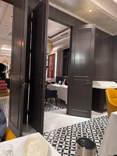 Captured indoors, the image showcases a unique perspective within a restaurant in Qinhuai District, China. The interior features dark-framed, divided doors that are either open or closed, revealing glimpses into different areas of the establishment. Patrons and staff can be seen through the openings, engaged in dining or service activities. The decor includes artwork, distinctive light fixtures, and patterned black and white tile flooring. The view is taken from a high angle. In the foreground, portions of a table setting are visible.