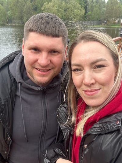 A selfie depicts a man and a woman outside near a body of water, possibly a lake. The woman, positioned at the top of the frame, has blonde hair and is wearing a red hooded sweatshirt under a black puffer jacket. The man, located below the woman, has short, dark hair and is wearing a gray hooded sweatshirt and a black jacket. The background features a body of water with trees on the opposite bank. The location is likely Grebnevo, Russia.