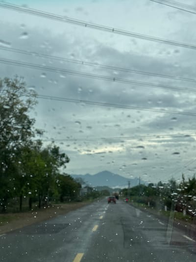 The image captures a perspective from inside a vehicle, looking out through a rain-streaked windshield at a rural road scene, likely in Thôn Thượng, Vietnam. The glass surface is covered with numerous raindrops, causing a soft-focus, blurred effect on the overall view.
The sky is heavily overcast with shades of grey and white clouds. Across the upper portion of the frame, several parallel lines, likely power lines or utility cables, stretch horizontally, partially obscured by the raindrops. Below these lines and extending into the midground, the silhouette of distant mountains or hills is visible against the cloudy backdrop.
To the right, a dark, wet asphalt road with a yellow dividing line extends into the distance. Green trees and foliage line the right side of the road, their forms somewhat blurred by the rain on the glass. In the mid-distance on the road, another vehicle, possibly a truck or bus, is visible, marked by its illuminated red taillights.
The weather is clearly rainy or has recently rained, evidenced by the drops on the glass and the wet appearance of the road. The time of day appears to be daytime, but the heavy cloud cover gives the scene a subdued, diffused light. No people are visible, and there is no discernible text in the image. The scene conveys a sense of travel through a natural landscape during inclement weather.