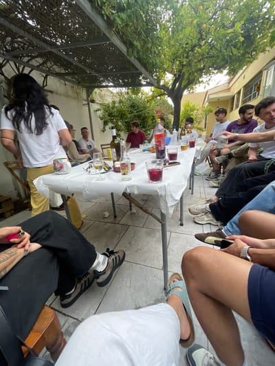 A group of people are gathered around a table outdoors. The table is covered with a white cloth and has various beverages and glasses on it. The people are dressed casually and appear to be socializing. The location appears to be a courtyard or patio in Jerez De La Frontera, Spain, with a tree providing shade and buildings visible in the background. There are at least 12 people gathered around the table with more potentially out of frame.