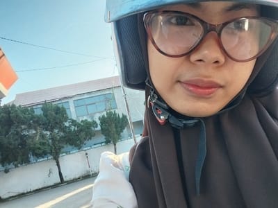 In this close-up, selfie-style photograph taken outdoors, a young woman is the primary subject. She is wearing a blue helmet and a brown hijab, partially obscuring her head and neck. Large, brown-rimmed glasses rest on her nose, and her face is turned towards the camera, looking slightly to the left. Her lips are colored with a reddish tint.

The background suggests an urban or semi-urban setting in Macege, Indonesia. Behind the woman, a building with a tiled roof and large windows can be seen. Several trees with dense green foliage stand in front of the building, casting shadows on a light-colored wall. The ground in the foreground appears to be a paved surface, possibly a road or sidewalk, with sunlight creating distinct shadows and highlights, indicating it is daytime. The sky is a clear, bright blue, suggesting fair weather.

A white fabric, possibly clothing or a scarf, is visible in the lower left of the frame. A strap from the helmet is also visible, dangling down the woman's right side. The overall mood of the image is casual and possibly indicative of travel or an outing, given the helmet. There is no visible text in the image.