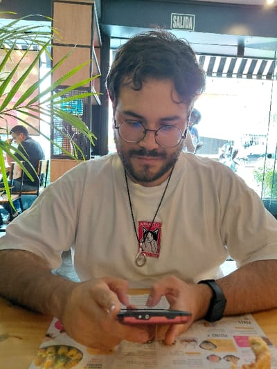 A person with glasses and a beard is sitting at a table, looking at a mobile phone. The person is wearing a white t-shirt with a graphic design and a black smartwatch. A menu is on the table next to the phone. There's a plant in the foreground and another person visible in the background. The scene appears to be inside a restaurant or cafe, with natural light coming from the outside.