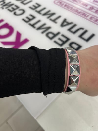 The image shows a close-up of a person's wrist wearing a pink bracelet with silver pyramid studs. The wrist is covered by the sleeve of a black garment. In the background, partially visible text suggests a location possibly related to retail or advertising, with some letters in purple and others in black. The text in the background appears to be in Russian and includes phrases like "БЕЗ ПЛАТИТЕ СЕЙЧАС" and possibly "3-6 МЕСЯЦ". The setting seems to be indoors, possibly a shop or a display area. There is no information in the image to suggest the location is Zanevka, Russia.