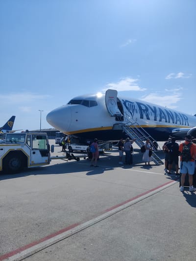 A Ryanair airplane is parked on an airport tarmac under a clear blue sky, suggesting a sunny day. A group of people are gathered near the open boarding stairs of the plane, indicating they are either boarding or disembarking. A small white tug vehicle is positioned in front of the airplane, possibly used for towing. In the background, another Ryanair plane's tail fin is visible, along with airport buildings and a truck. The scene appears to be at an airport, with the presence of airplanes, tarmac, and ground support equipment. The time of day is likely daytime due to the bright sunlight and clear sky. There is no explicit indication of emotions, but the activity suggests a normal travel day. The visible text on the airplane reads "RYANAIR". There is also text on the tug vehicle that is partially visible, appearing to say "SERVEFLY". The location is identified as Weeze, Germany.