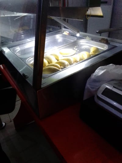 The image shows a stainless steel food warmer with a glass lid, located at latitude 5.936, longitude 5.657 in Sapele, Nigeria. The warmer is partially open, revealing several baked pastries, presumably pastries. To the right of the warmer is a cash register, sitting on a red table. A white plastic bag is next to the cash register. The scene is lit by artificial overhead lights. There are no people in the image.