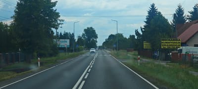 The image shows a straight, paved road with white dashed lines dividing the lanes. The road is bordered by grassy areas and trees on both sides. There are streetlights and power lines visible overhead.

On the left side of the road, there is a billboard that appears to advertise a business. Further down the road on the left, there is a black metal fence.

On the right side of the road, there are several yellow billboards advertising "MECHANIKA KLIMATYZACJA WULKANIZACJA" and a phone number. A white van with "Crena" written on it is parked near these billboards, and a portion of what appears to be a tire is visible. There is also a residential building with a tiled roof.

Several cars are visible on the road, moving away from the camera. One white car is in the foreground. There are also road signs, including a pedestrian crossing sign. The sky is cloudy.

The scene depicts a road in a semi-rural or suburban area, likely in Wola Filipowska, Poland, given the Polish text on the billboards and the overall environment. There are no people clearly visible in the image. The overall impression is of a typical roadside scene with commercial advertisements and passing traffic.