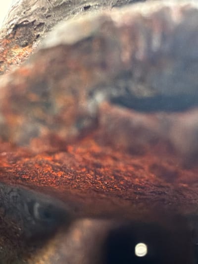 This is a macro photograph of a rough, textured surface, possibly wood or a rock formation with visible signs of decay or weathering. The dominant colors are earthy browns, oranges, and grays, with areas of intense reddish-orange suggesting rust or intense oxidation. The surface is irregular with crevices and a porous texture. In the lower portion of the image, a blurred, circular, white light source is visible, possibly a reflection or a distant light. The focus is on the intricate details of the textured material, creating an abstract composition. The image does not contain any recognizable elements of the city of Grapevine, United States of America, nor does it depict any people, subjects, or actions. The environment appears to be natural or weathered, without any urban context.