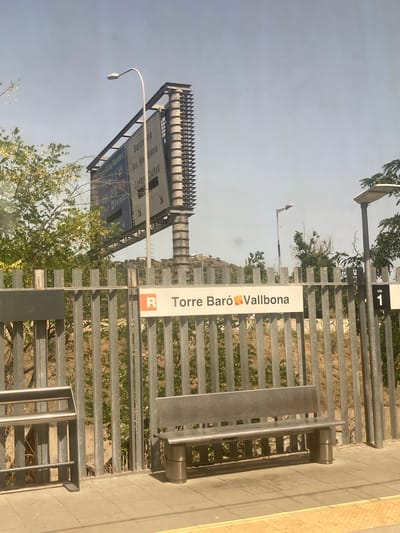 The image captures a section of the Montcada i Reixac train station in Spain. In the foreground, a metallic fence runs horizontally across the frame, separating the platform from the tracks. A modern, metallic bench is situated on the platform. Above the fence, a station sign reads "R Torre Baró Vallbona" with a small orange and yellow emblem to the left of "Vallbona". To the right of the sign, a black square indicates "via 1". In the background, a large billboard with directional information for various destinations, including "Barcelona" and "Aeroport", is visible. Green foliage and trees are present behind the fence and billboard. The sky is clear and light blue.