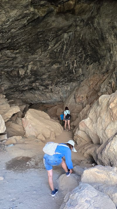 The image depicts the interior of a cave, with rough, textured rock formations dominating the scene. In the foreground, a person wearing denim shorts, a blue long-sleeved shirt, a white patterned backpack, and a white wide-brimmed hat is bent over, adjusting their dark sneakers on a rock. Further into the cave, two other individuals are visible. One, wearing a light blue shirt and dark shorts, stands facing away from the camera with a black backpack. Next to them, a child in a blue shirt is also facing away. The cave floor is composed of dirt and gravel, with several large boulders scattered throughout. The cave's ceiling is a mass of dark, uneven rock. The location is identified as Terekty, Kazakhstan.