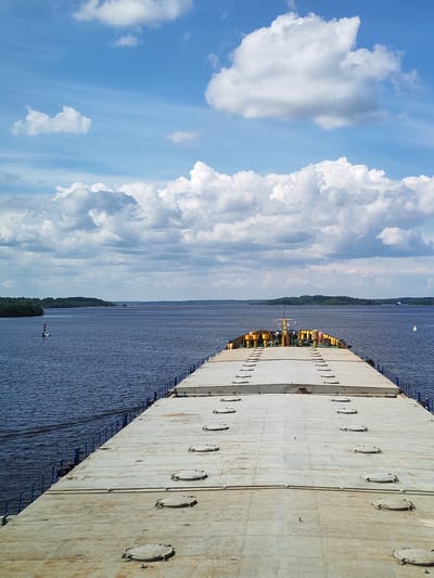 The image presents a view from the deck of a barge on a wide body of water, with land visible in the distance. The barge's deck is a flat, gray surface with numerous round hatches. Blue railings line the edges of the deck. In the distance, a small control tower is visible on the barge. The water is dark, with small waves. On the left side of the frame, a navigational buoy is visible. The sky is blue with scattered, puffy white clouds. Based on the prompt, this scene is in the city of Zvoz, Russia. The image captures a scene of maritime transport.