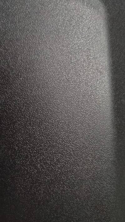 The image displays a close-up, textured surface with a dark gray, matte finish. The texture is characterized by a fine, irregular pattern of raised ridges and indentations, creating a somewhat mottled appearance. The lighting creates subtle shadows and highlights that emphasize the three-dimensionality of the surface. There is a gradual transition to a lighter gray area on the right side of the image, suggesting a curved surface or a change in material. No people, specific objects, actions, or environmental context are visible. The image is purely a macro shot of a textured material.