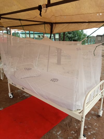 A metal hospital bed with wheels is set up outdoors under a makeshift canopy. The bed is covered with a white mosquito net, which drapes over the frame and down to the sides. A white pillow with a subtle pattern rests on the bed. The mattress and pillow are also covered by the net. A red rug is placed on the ground in front of the bed. The ground appears to be dirt or sand. In the background, there are some trees, buildings, and a fence. The sky is visible through the canopy, suggesting it is daytime. There are some labels on the bed frame, one of which is partially visible and reads "MCRP" followed by other text. The overall setting appears to be a simple, outdoor medical or temporary shelter area in Yola, Nigeria.