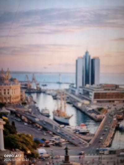 The image is a blurry, telephoto shot of a city harbor at sunset. In the foreground, a Ukrainian flag is visible on a building. A large, modern skyscraper dominates the background, with several boats and cranes in the middle ground, suggesting a busy port. The sky is filled with soft, pastel-colored clouds. The timestamp "2026/07/26 22:01" is superimposed on the lower left corner. The location is identified as Құмөзек, Kazakhstan, however, the architecture and the presence of the Ukrainian flag do not align with this identification. The scene depicts a coastal urban area with significant maritime activity.