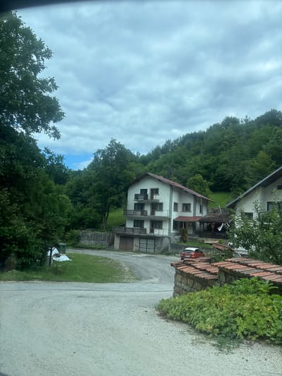 The image depicts a rural scene in the city of Shipkovo, Bulgaria, characterized by two residential buildings nestled amidst lush greenery and a backdrop of forested hills under a cloudy sky.
The foreground features a gravel driveway leading towards the buildings. To the right, a low stone wall topped with terracotta roof tiles separates the driveway from an area with dense green foliage.
The main building, on the left, is a multi-story house with a white facade, prominent balconies with railings, and a red-tiled roof. It has multiple windows and what appear to be garage doors at the ground level. A red and white car is parked in front of this building. To its right, another residential building, also white with a red-tiled roof, is partially visible. It also has balconies and windows. A covered outdoor area with a tiled roof is situated between the two buildings, adorned with a flag featuring red, white, and green bands.
The environment is predominantly natural, with trees and vegetation surrounding the structures. The hills in the background are densely covered with trees. The sky is overcast, with patches of lighter clouds suggesting diffused daylight. A green refuse bin and some covered items are visible on the grass to the left of the driveway. The overall impression is of a quiet, somewhat secluded residential area within a natural landscape.