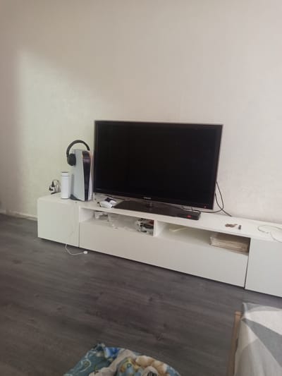This is a medium-range, eye-level, indoor, full-color, slightly grainy photograph taken in a room in Hlusk, Belarus. The scene depicts a modern living room setup with a white entertainment stand against a pale, textured wall. On the left side of the stand, a white PlayStation 5 console stands vertically, topped with a pair of black headphones. To the left of the console is a white cylindrical object, possibly a water bottle or speaker, with a black charging cable plugged into a power adapter behind it.
The entertainment stand features a long, white surface with shelving. The television, a large Samsung flat-screen, is centrally placed on the stand. Below the television, on the shelf, are two white game controllers. To the right of the controllers, on the same shelf, are several items, including what appears to be a small black electronic device with red indicator lights, a stack of papers or magazines, and some loose change. A white charging cable is visible on the floor in front of the stand, connected to a white circular object.
The floor is covered with dark gray laminate flooring, with visible wood grain. In the foreground, a portion of a colorful blanket or bedding is visible. The overall impression is that of a tidy, contemporary living space.