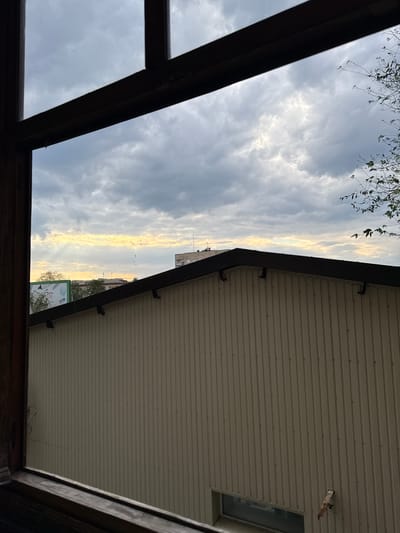 The image presents a view through a wooden window frame, looking outwards at a partly cloudy sky and urban rooftops. The sky exhibits dramatic clouds with streaks of sunlight breaking through, creating a dramatic contrast between light and shadow. Below the sky, a sloped, dark-roofed structure with light-colored, ribbed siding dominates the mid-ground. In the distance, beyond this structure, are glimpses of other buildings and a billboard. The scene appears to be set in Stukalova Balka, Ukraine. There are no people or animals visible in the image. The overall impression is of a somewhat overcast day in an urban or semi-urban environment.