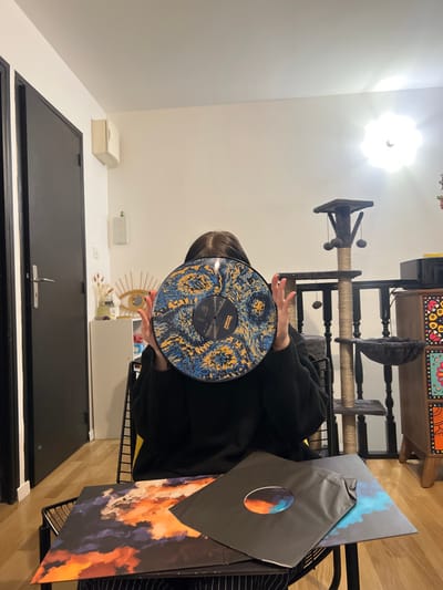 A person in a black sweater holds up a colorful vinyl record, obscuring their face. The record has a complex pattern of blue, black, and gold. To the right, the vinyl record packaging features a cloud-like, abstract design in shades of orange and blue. Background details include a black door, a cat tree, and a wooden cabinet. The room is well-lit.