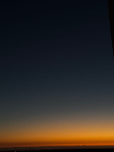 The image shows a view of Earth from space, capturing the curvature and the atmospheric transition from dark blue to orange. Two faint points of light are visible in the dark blue portion, likely stars or planets. Along the horizon, a scattering of brighter points of light suggests the presence of a city or settlement at night. There is no verifiable data in the image to identify the city as Ariel, Palestinian Territories.