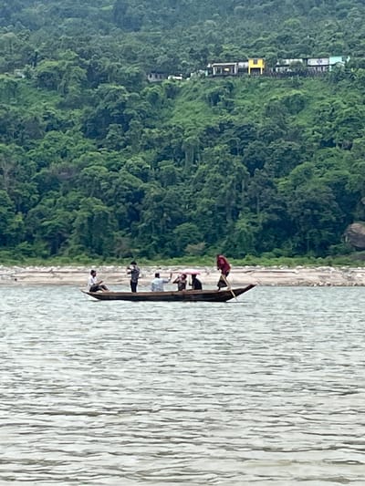 A wooden boat with several people is on the water in front of a rocky shore and a densely forested hill with scattered houses. The boat appears to be a shallow, elongated vessel, possibly used for transportation or fishing. There are at least five individuals visible in the boat. One person, wearing a dark red shirt, stands at the stern and appears to be poling the boat with a long stick. To their left, another person is seated. Further forward, three other individuals are clustered. One of them is holding a pink umbrella. The water has a rippled surface, reflecting the overcast sky. The background consists of a steep hillside covered in lush green trees. The houses on the hillside are of varying colors, including yellow, white, and light blue. The overall scene suggests a natural landscape with signs of human habitation. This is likely the Jaflong region of Bangladesh, known for its rivers and hilly terrain.