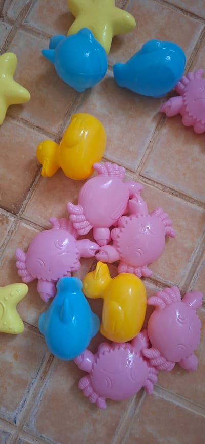The image displays a collection of brightly colored plastic toys, likely designed for beach or sand play, scattered across a tiled surface. The subjects are the toys themselves, which include several pink crab-shaped molds, yellow duck-shaped molds, blue dolphin-shaped molds, and yellow star-shaped molds. These objects are arranged in a somewhat haphazard fashion, suggesting they have been used or are about to be used.
The scene is indoors, given the tiled floor which appears to be a common flooring material in bathrooms or kitchens, not typically found outdoors in a beach setting. The lighting is uniform, suggesting indoor lighting, and there are no indicators of weather conditions or time of day. The overall emotion evoked by the image is playful and perhaps nostalgic, due to the nature of the toys.
There is no visible text in the image. The location context of Orimattila, Finland is provided externally and does not manifest in any visual cues within the image itself. The activity or event taking place is implied to be play or storage of these toys. The tiled surface has a slightly worn appearance with some grime in the grout lines, adding a subtle detail to the setting.