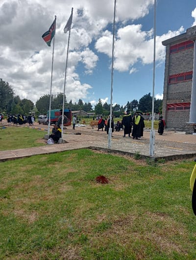 The image portrays an outdoor institutional area in Waseges ward, Kenya, under a partly cloudy sky. In the foreground, green grass with dry patches gives way to a paved walkway and a larger paved area. A small reddish-brown soil mound is present in the grass.
Four vertical flagpoles stand in the midground; the leftmost displays the Kenyan national flag, the second flies a plain grey flag, and the remaining two are unadorned. Numerous individuals are visible across the paved area, with a prominent group wearing black academic gowns, some with yellow or green stoles/hoods, suggesting a graduation event. These individuals are standing, walking, and interacting.
To the left midground, a blue and green cargo truck is parked. Near the truck, several individuals are engaged in activities involving wooden structures, possibly setup or dismantling, with one person using an umbrella for shade. A person in a black gown is seated on a low wall or step to the left, with a light-colored bag beside them.
The background features a multi-story building constructed with stone blocks and red-framed elements on its upper levels, situated on the right side of the frame. A line of trees forms the distant green backdrop beneath a blue sky adorned with white cumulus clouds.