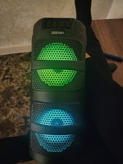 A close-up, high-angle shot shows a black ZQS2401 portable speaker with LED lights. The speaker is upright and is illuminated with green lights on the top half and blue lights on the bottom half. The speaker sits on a dark surface, with a wood-grain surface visible to the right, and a patterned wall or carpet to the left. A dark-colored sock or fabric is visible behind the speaker. A blue pen lies on the wood-grain surface to the right of the speaker. The image is dark, with most of the background obscured by shadow. There is no visible indication of the city of Gudermes, Russia, or any other specific location.
