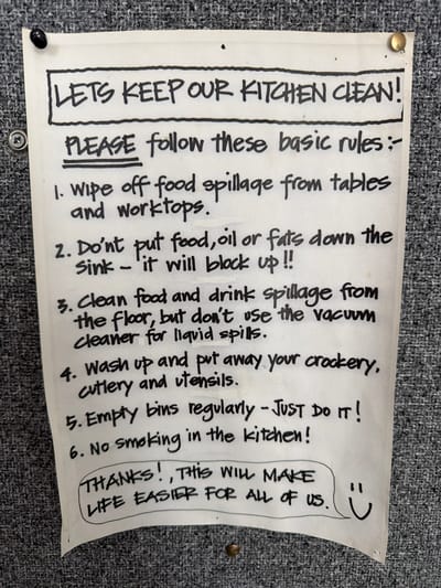 A handwritten note is pinned to a textured grey wall. The note, written in black marker, lists six rules for keeping a kitchen clean. The title, in large block letters, reads "LETS KEEP OUR KITCHEN CLEAN!". Below this, in smaller but still prominent letters, is "PLEASE follow these basic rules:". The rules are numbered and begin with "1. Wipe off food spillage from tables and worktops." and continue with instructions to avoid putting food, oil, or fats down the sink, to clean spills from the floor without using a vacuum cleaner, to wash up and put away crockery and utensils, to empty bins regularly, and a prohibition against smoking in the kitchen. The note concludes with a circled message that reads "THANKS! THIS WILL MAKE LIFE EASIER FOR ALL OF US." and a smiley face. The note appears to be in a communal area, possibly an office or staff kitchen. There is no information in the image that places it specifically in Epsom and Ewell, United Kingdom.