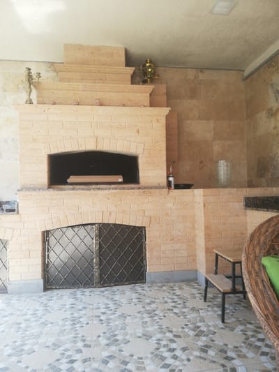 The image depicts an outdoor kitchen area, likely a patio, featuring a prominent brick oven. The oven is constructed from light-colored bricks and has a large arched opening. Inside the oven, a long, flat, rectangular object wrapped in cardboard is visible. Below the oven, a metal grate door covers a storage area. To the right of the oven, a small table with wooden benches and a woven chair with a green cushion are situated. The floor is tiled with a decorative pattern of light and dark geometric shapes. In the background, a wall is tiled with large, irregularly shaped stone tiles. On top of the oven structure, there are decorative elements, including a brass samovar and a brass candelabra. A small butane torch and a dark, shallow pan are placed on a ledge to the right of the oven opening. The overall setting suggests a functional and somewhat decorative outdoor cooking or gathering space. The scene is consistent with an outdoor area in a residential or public space in Akhalkalaki, Georgia, given its architectural style and typical outdoor kitchen setups found in the region.