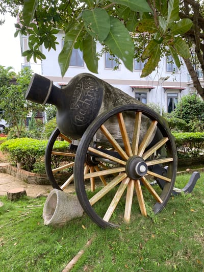 The image depicts an old cannon with large wooden wheels, positioned on a grassy area with some vegetation. A building with multiple windows and red-tiled roofs is visible in the background, suggesting a residential or commercial setting. The cannon itself appears to be made of dark material and has the inscription "Puch Backmann" in a circular pattern on its barrel. The time of day appears to be daytime, with natural light illuminating the scene. The weather seems fair. The location is identified as Thôn 2, Vietnam. No people are visible in the image. The overall scene evokes a sense of history and perhaps a decorative display of an antique object.