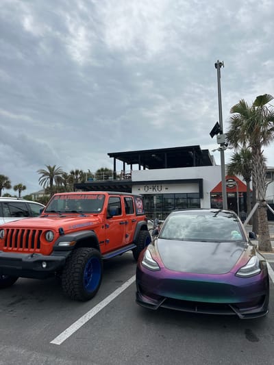 The image shows two cars parked outside the restaurant "O-Ku" in Jacksonville Beach, Florida, United States of America. The sky is overcast. There is a white car, an orange Jeep with a "GATORLATION" decal, and a color-shifting (possibly iridescent) Tesla Model 3. The restaurant has a black awning and glass doors, with outdoor seating visible on a patio. A palm tree is visible next to the building. A pole with a camera and light fixture is visible above the cars. "UKUMADES" is written on an orange sign next to the restaurant.