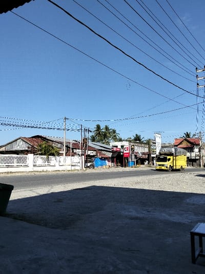 A sunny street scene features a yellow truck driving on the road. Buildings with rusty roofs line the street, interspersed with palm trees and utility poles with many hanging wires. A white fence runs along the left side of the frame. The foreground is a shaded area, partially obscuring the view, with glimpses of a table and trash can. The sky is a clear blue.