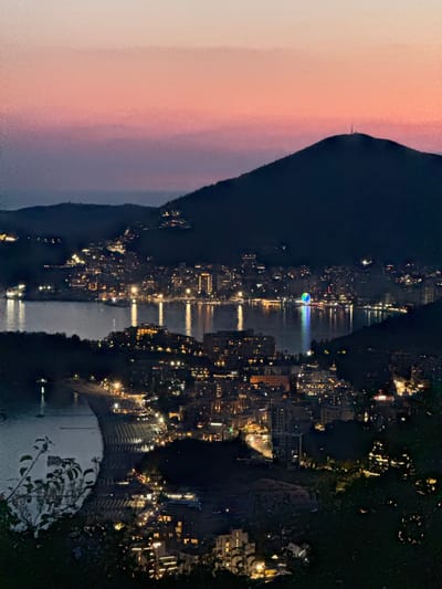 This is a nighttime, aerial, long-shot photograph of the city of Podgorica, Montenegro, as dusk transitions to night. The scene is dominated by a panoramic view of the city lights spread across a coastal area, with dark, silhouetted mountains in the background. The sky displays a gradient of twilight colors, transitioning from a pale pink at the top to a deep purple near the horizon.
The city is illuminated by a multitude of artificial lights, creating a vibrant urban landscape. Buildings, ranging from hotels to residential structures, are clearly visible, their windows glowing warmly. A body of water, likely the sea or a large bay, is present in the foreground, reflecting the city lights. A noticeable feature in the water is a Ferris wheel, brightly lit with blue and green lights, standing out against the darker surroundings.
The coastline features a wide, crescent-shaped beach lined with numerous beach chairs and umbrellas, though they are difficult to discern clearly in the low light. The beach is also bordered by buildings and what appears to be a road or promenade. The overall impression is one of a lively, illuminated city at the edge of the sea, set against a dramatic mountainous backdrop during twilight. The photograph is taken from a high vantage point, offering a comprehensive view of the urban sprawl and its natural environment.