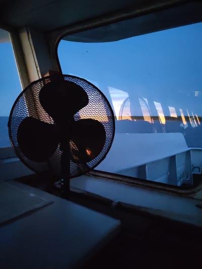 The image shows a metal mesh fan placed on a white surface, presumably a table or shelf, inside a room with a window. Outside the window, a body of water (likely a river or lake) can be seen, with a strip of land or coastline visible in the distance. The lighting suggests it is either dawn or dusk. The fan's reflection is visible in the window. The structure framing the window suggests the scene might be inside a boat or a building near water. The image does not provide enough information to determine the specific location in Kostyaevka, Russia.