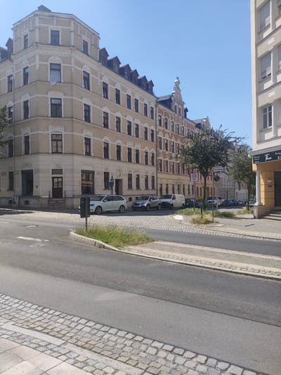 The image captures a street scene in Chemnitz, Germany, on a bright, sunny day. The foreground shows a paved road with a cobblestone sidewalk and a pedestrian crossing. In the midground, several cars are parked along the street, including a white station wagon and a dark sedan. A white van is also visible. Behind the cars, a row of multi-story buildings with light-colored facades lines the street. The buildings feature numerous windows and decorative architectural details.
One building on the left has a prominent corner entrance with stairs leading up to it. Another building on the right displays a sign with the text "Friseur Kosmetik" and "TRAUM," indicating a hair salon or beauty business. A speed limit sign for 30 km/h is also visible, along with a street sign that reads "Tiefenstraße." The scene suggests a typical urban environment during daylight hours, with clear blue skies overhead. The overall impression is one of a quiet, residential or commercial street on a pleasant day.