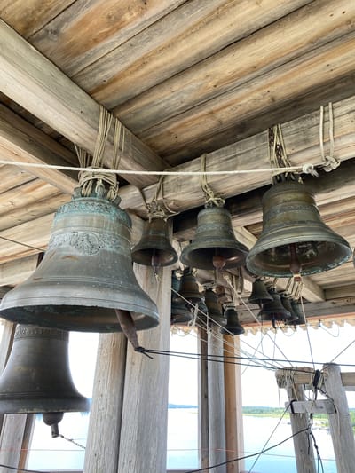 In Boyarshchina, Russia, a collection of large bronze bells are suspended from a wooden structure. The bells vary in size and are adorned with decorative bands and some with inscribed text. They hang from thick ropes tied to wooden beams, which form the roof of the structure. The wooden beams show signs of age and weathering. In the background, a body of water can be seen, with a distant shoreline dotted with trees. The overall scene suggests a bell tower or a belfry.
