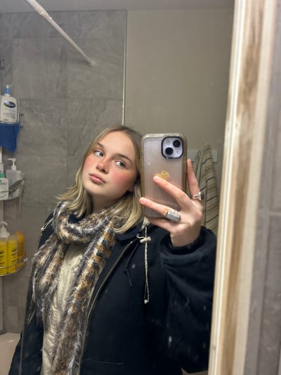 A young woman poses for a mirror selfie in a bathroom. She wears a dark jacket with a faux fur-trimmed scarf. She holds an iPhone with a clear case adorned with a dandelion graphic. She sports silver rings on her right hand and has a fair complexion with rosy cheeks. The bathroom features grey tiles and various toiletries are visible on a shelf. The lighting is bright and somewhat unflattering.