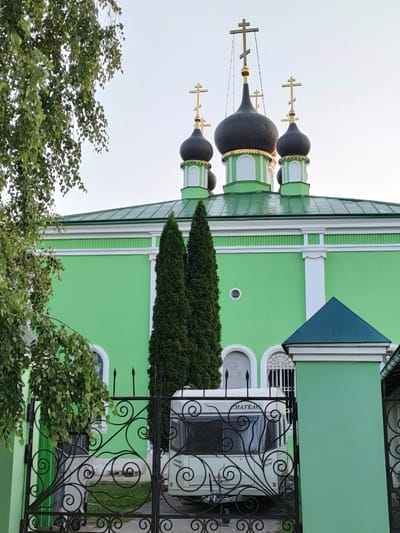 This is a medium-shot, eye-level, outdoor photograph of a bright green church with multiple black onion domes topped with golden crosses. The church has a green roof and white decorative trim along its upper section. In front of the church, there is a black wrought-iron gate with ornate scrollwork. Through the gate, a white caravan is partially visible. Two tall, slender evergreen trees stand to the right of the gate, framing the caravan and the church. To the left of the frame, a leafy green tree branch with small leaves hangs down. The sky is a pale, overcast color, suggesting it is either early morning or late afternoon. The overall scene depicts a tranquil, somewhat pastoral setting. There is some visible text on the caravan that reads "CHATEAU". The location context provided is Boltino, Russia.