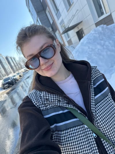 A young woman is depicted from the chest up in a bright outdoor selfie, smiling faintly directly at the camera. She wears brown oversized sunglasses with light blue tinted lenses. Her attire consists of a light pink t-shirt, a dark brown hooded jacket, and an outer garment featuring a black and white houndstooth pattern with horizontal black and light grey stripes across the chest. A dark green strap, likely from a bag, crosses her chest. Her light brown hair is visible, pulled back. The background features a clear blue sky in the upper left. Multi-story residential buildings with light-colored facades and dark windows are visible on both sides of the frame. A street with parked vehicles, including a dark SUV, runs in the mid-ground, partially covered by melting snow and slush. Large snow piles are prominent on the right side, adjacent to a building entrance with a dark overhang. The scene is brightly illuminated by direct sunlight. The location is Новое Шигалеево, Russia.
